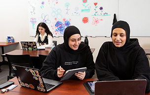 Two students work together on laptops while one student in the background works on her laptop.