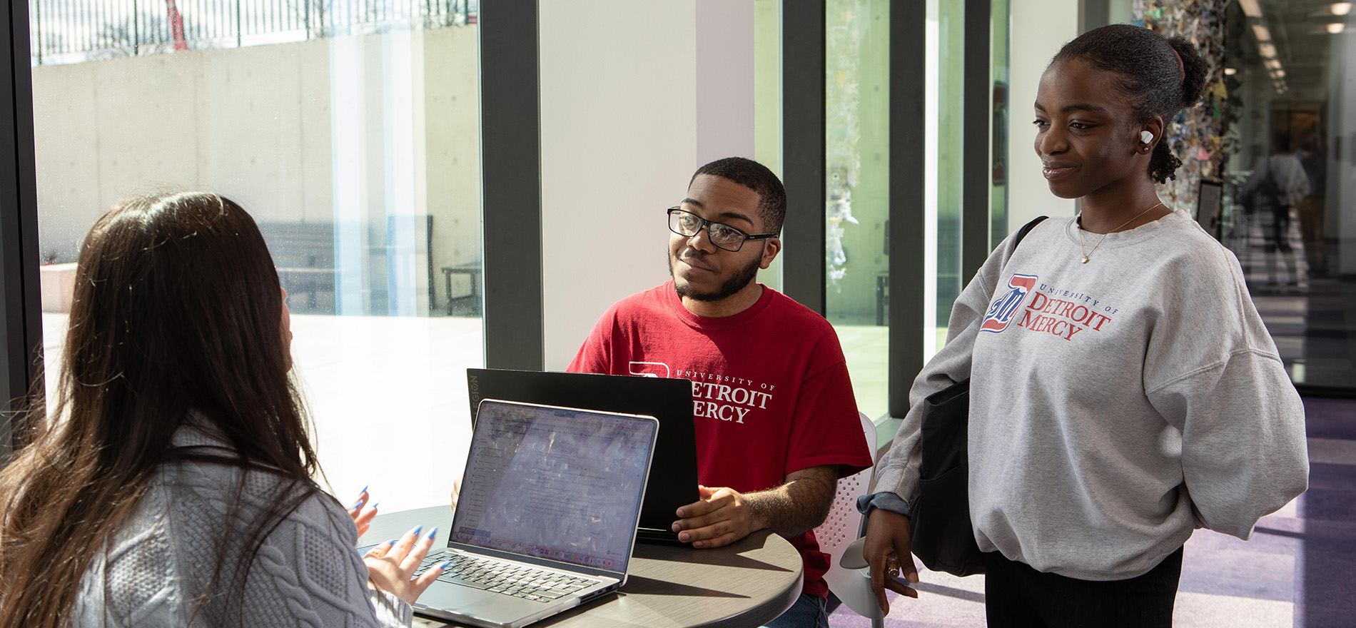 A group of students gather around a table with their laptops, while one stands.