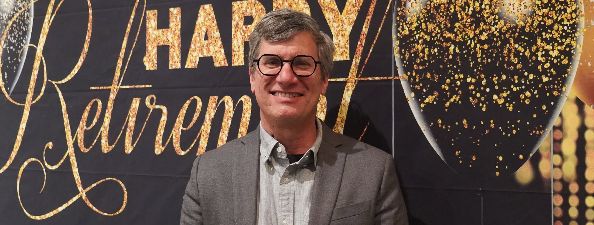 Nicholas Rombes standing and smiling in front of a Happy Retirement banner indoors.