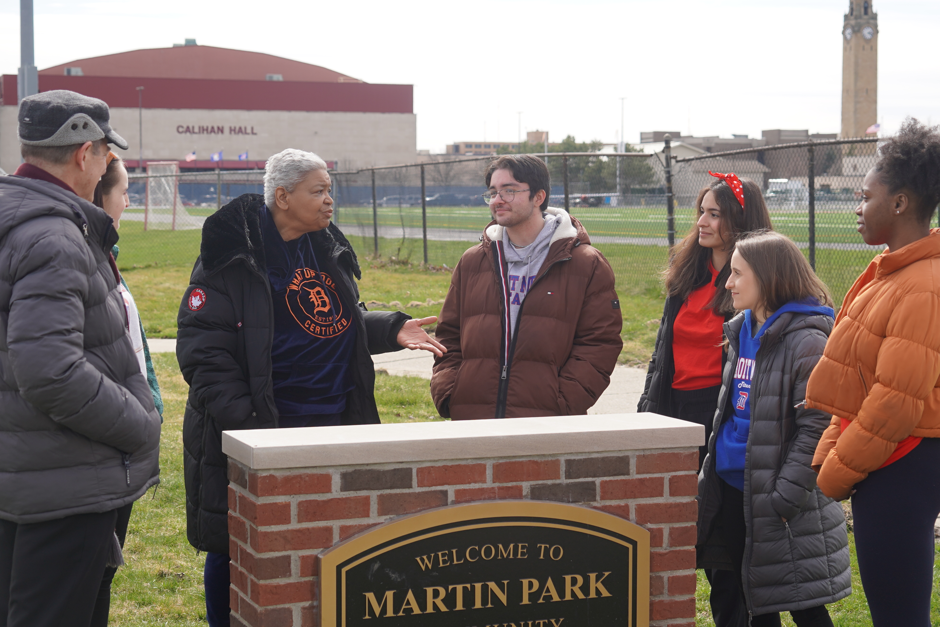 Seven people stand outdoors near a sign for Martin Park and the perimeter of the McNichols Campus.