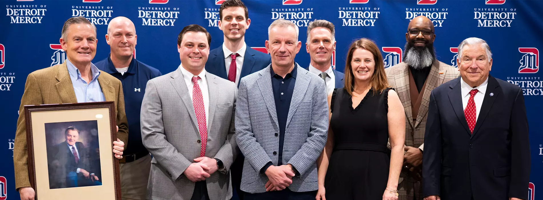 Nine members of the Hall of Fame are dressed up and standing in front of a blue University of Detroit Mercy backdrop.