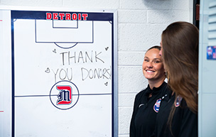 A women's soccer student-athlete smiles in the corner of a photo of a whiteboard that says Thank you donors <3