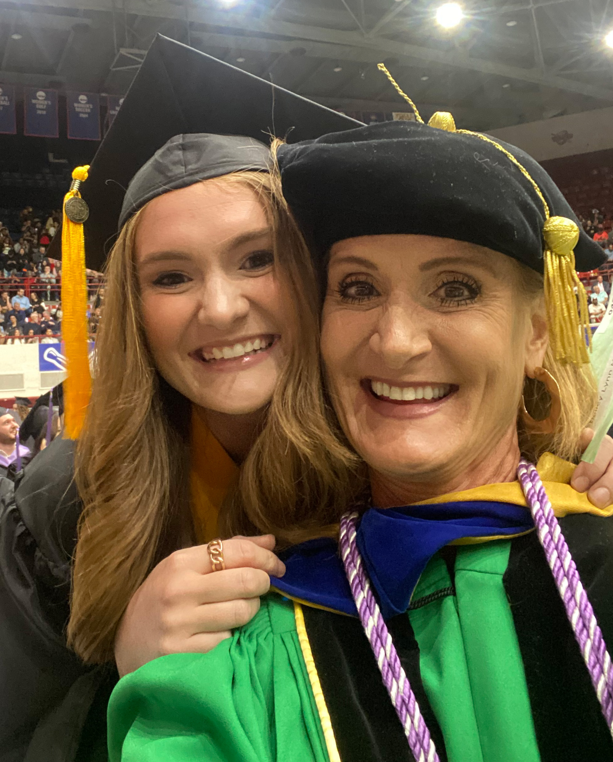 Two women smile for a photo during Commencement inside of Calihan Hall, with others in the background.