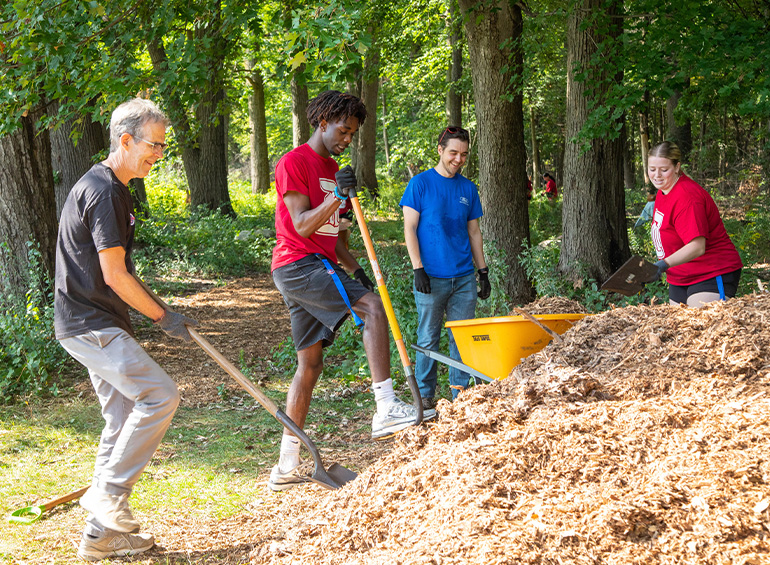 Students and two men work together by digging at a mulch pile.