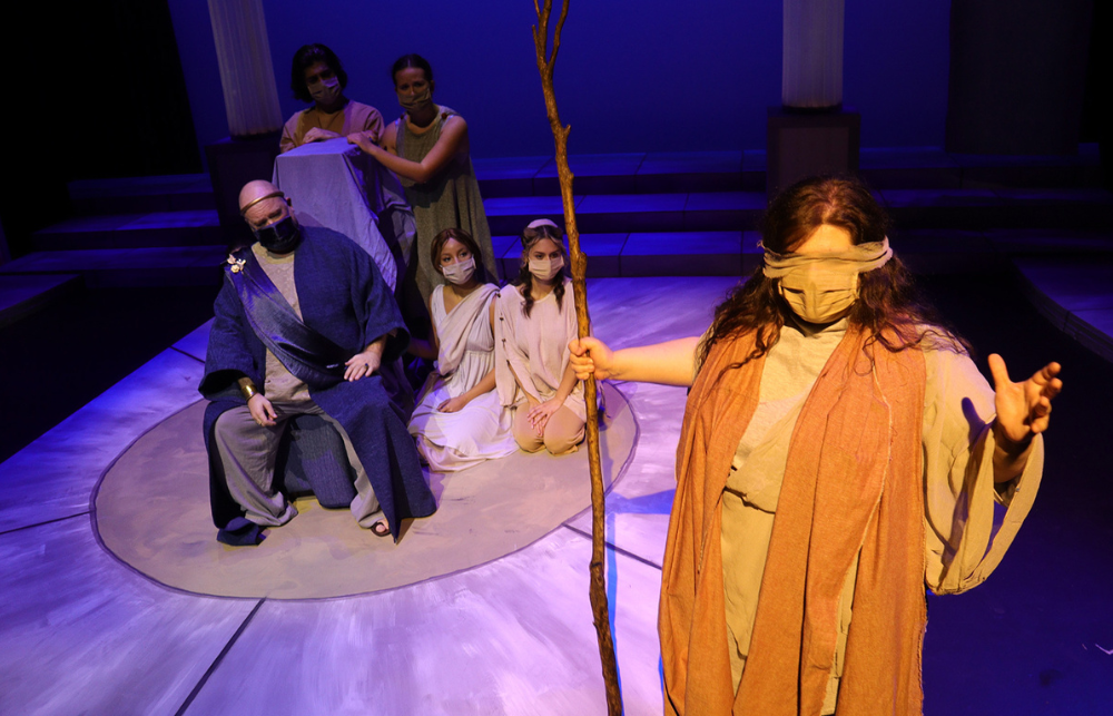 Six actors, wearing masks, perform indoors during the production of Antigone. The woman in the front of the photo is blindfolded and holding a staff stick.