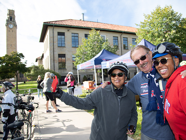 Three people pose for a photo wearing bike helmets, with other bikes and riders behind and campus buildings, trees and a clock tower beyond on a sunny day.