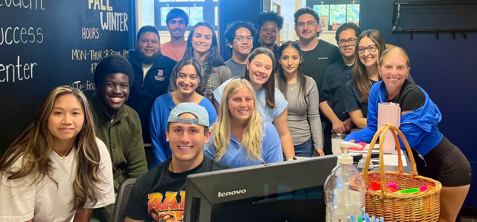 Eleven people, most standing, smile for a group photo inside of the Student Success Center. A blackboard reads, Testing 9 a.m. with SSC hours.