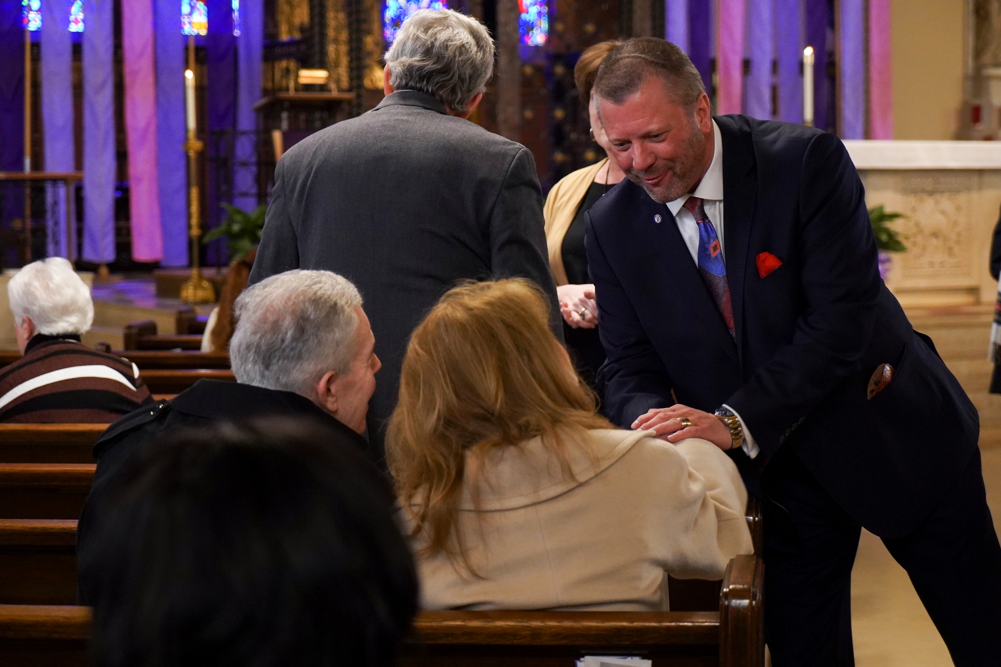 President Donald Taylor stands inside of a church, shaking hands with a pair of people sitting in a pew. Others are pictured in the background.