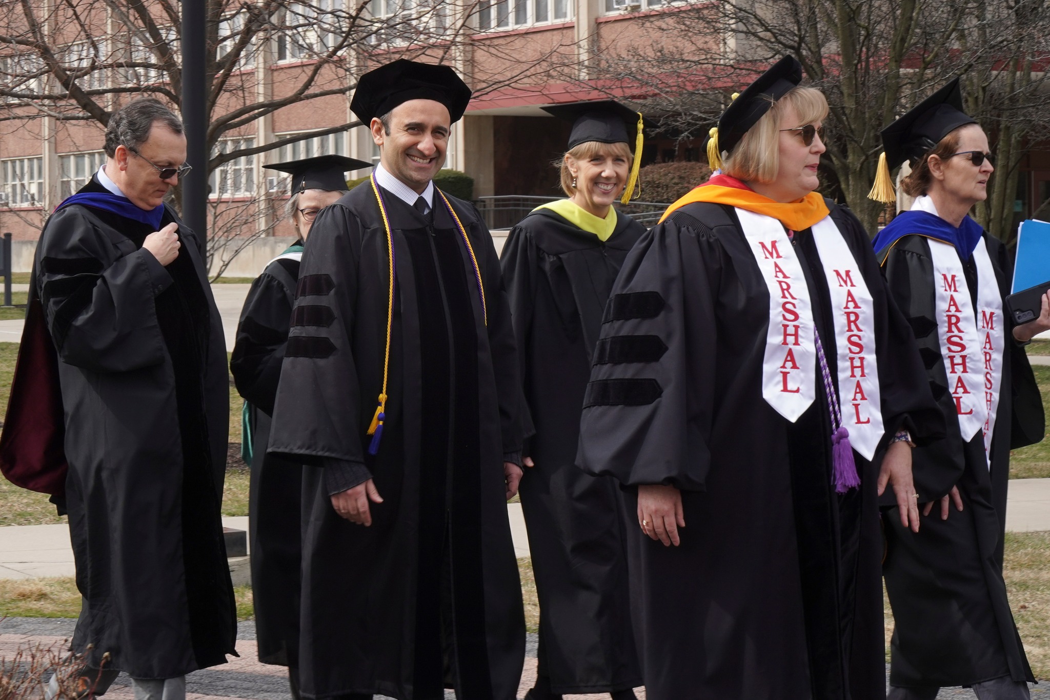 Six people walk outdoors, wearing robes, with two people wearing Marshal ribbons over their shoulders. Buildings, trees are pictured in the background.