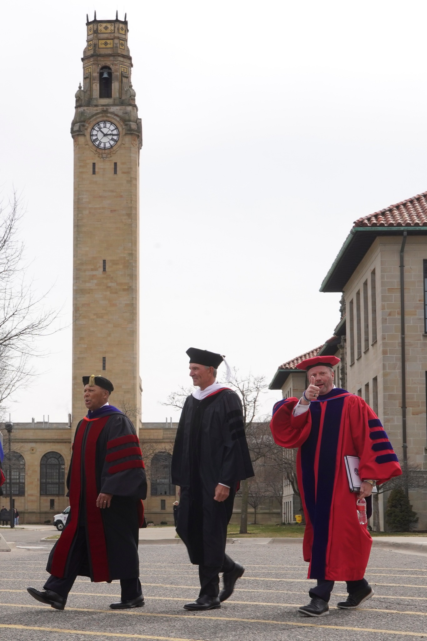 Three people walk on the sidewalk with buildings, trees and a large clock tower in the background.