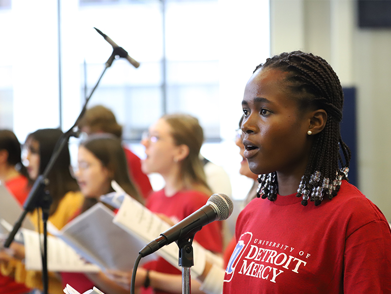 A student wearing a red University of Detroit Mercy t-shirt sings near a microphone with other students singing in the background.