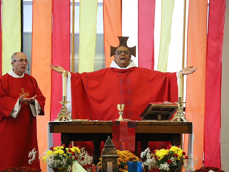 Two fathers stand on a stage, one holding open his arms in front of a pulpit.