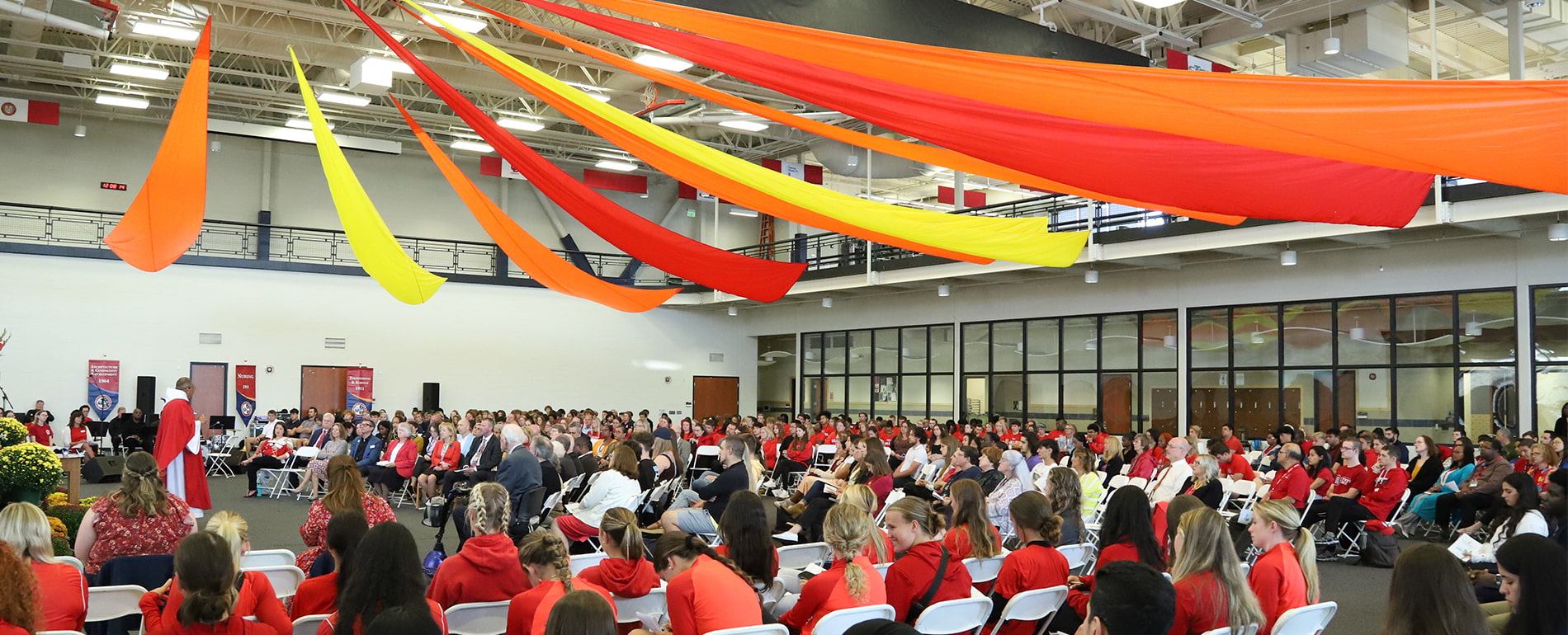 Dozens of people, in reds and orange and other colors, sit and listen during Celebrate Spirit inside of the Student Fitness Center. Large orange, red and yellow ribbons hang above.
