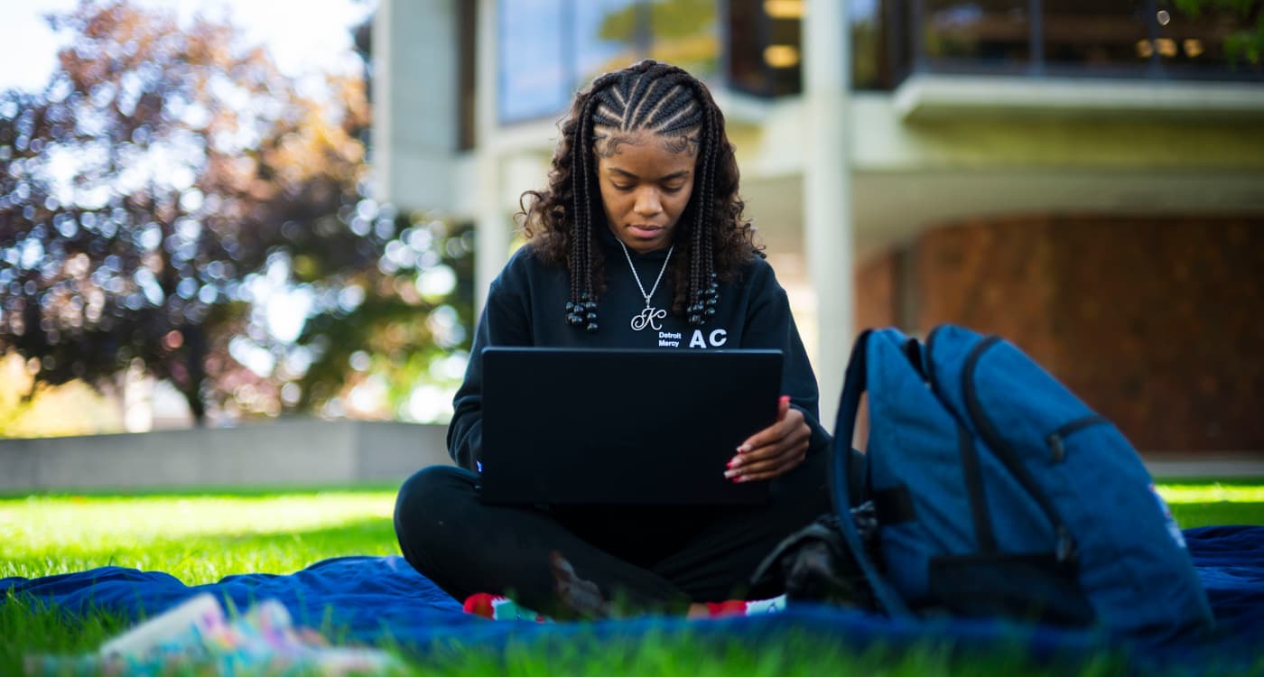 A student types on a laptop while sitting on a blanket outside.